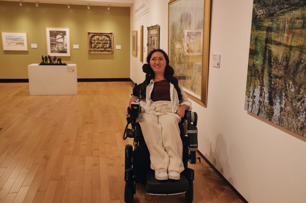 Tori Hunter, a wheelchair user and disabled travel blogger, smiling in her power wheelchair in a spacious, wheelchair accessible exhibition room at the Woodstock Art Gallery in Woodstock, Oxford County, Ontario