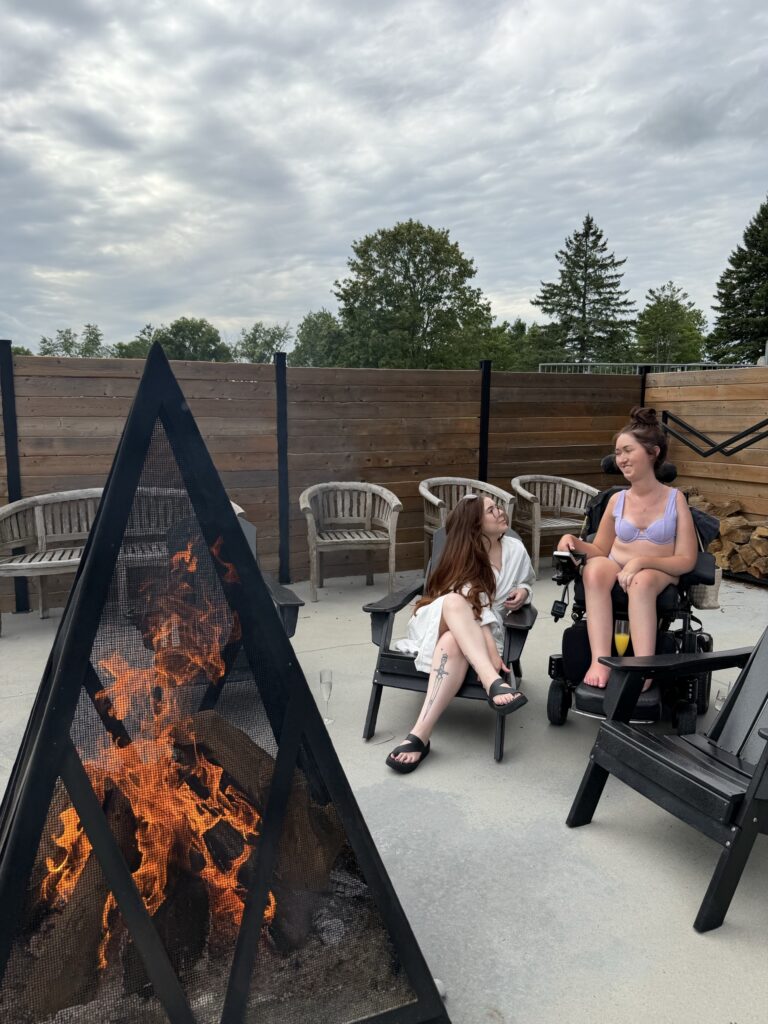 Tori Hunter, a wheelchair user and disabled travel blogger, chatting with a friend in her power wheelchair by the outdoor fire pit on the step-free concrete patio at Wave Nordic Spa in Mount Elgin, Oxford County, Ontario