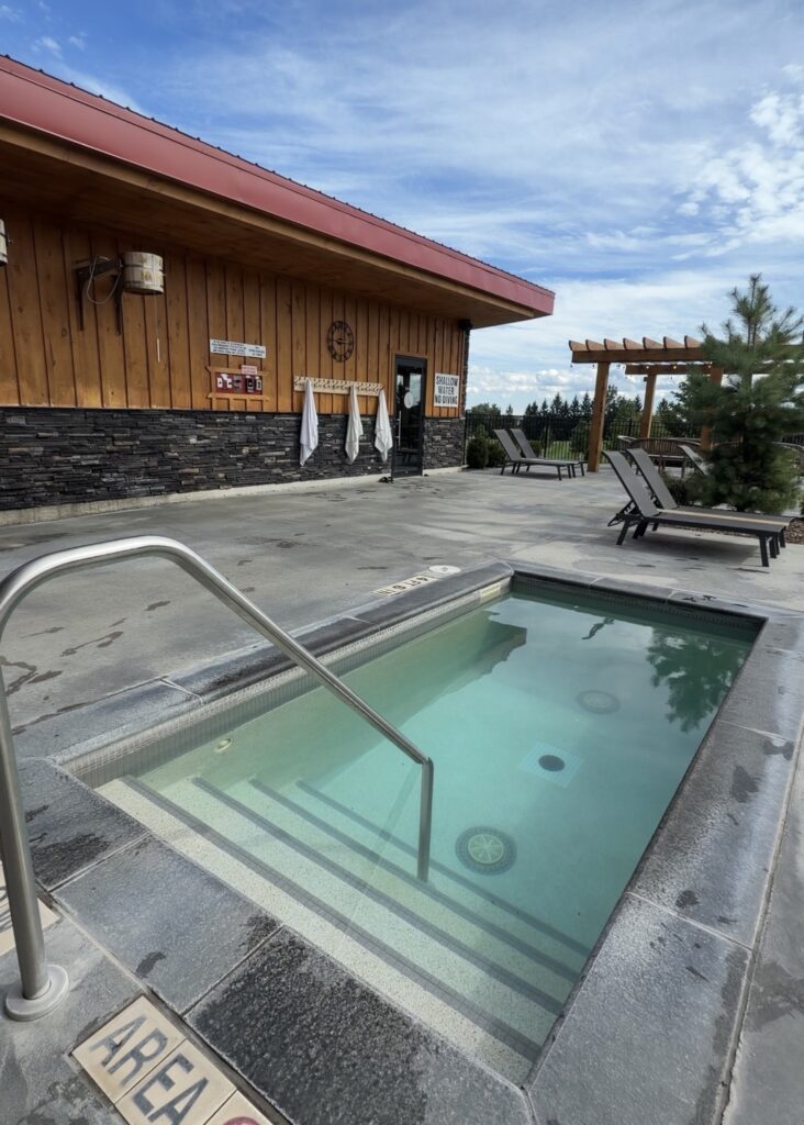 Outdoor hot pool with handrail and wide entry steps beside the wood-clad sauna building at Wave Nordic Spa, a wheelchair accessible Nordic spa at The Oxford Hills in Mount Elgin, Oxford County, Ontario