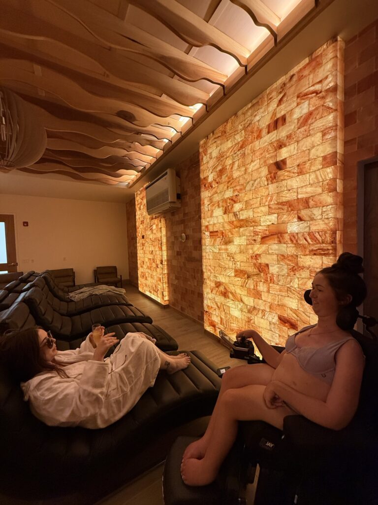 Tori Hunter, a wheelchair user and disabled travel blogger, relaxing in her power wheelchair beside a friend on a lounger in the glowing Himalayan salt room at Wave Nordic Spa in Mount Elgin, Oxford County, Ontario