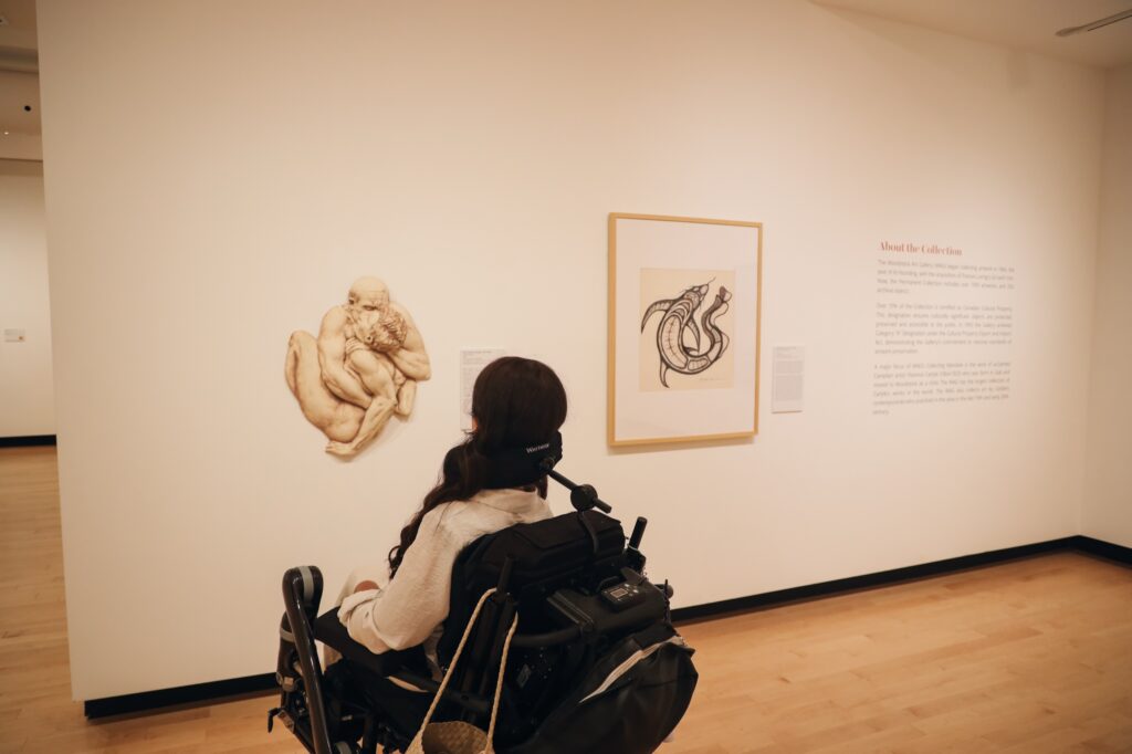 Tori Hunter, a wheelchair user and disabled travel blogger, viewing a sculpture and framed artwork from her power wheelchair in a wide, step-free gallery at the Woodstock Art Gallery in Woodstock, Oxford County, Ontario