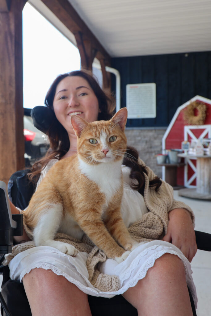 Tori Hunter, a wheelchair user and disabled travel blogger, smiling in her power wheelchair with a friendly orange and white cat on her lap on the step-free porch of Makkink's Flower Farm in Tillsonburg, Oxford County, Ontario