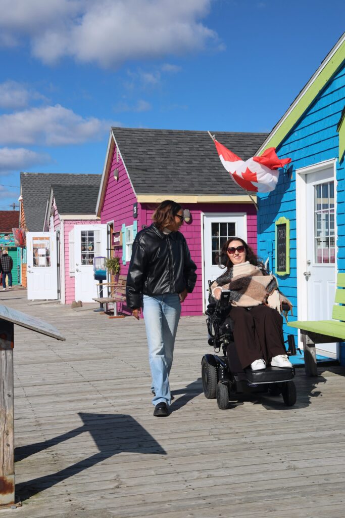 Tori Hunter, a wheelchair user and disabled travel blogger, in her power wheelchair with a friend on the boardwalk in front of the colourful pink, blue and green artisan shops at Fisherman's Cove in Eastern Passage, Nova Scotia