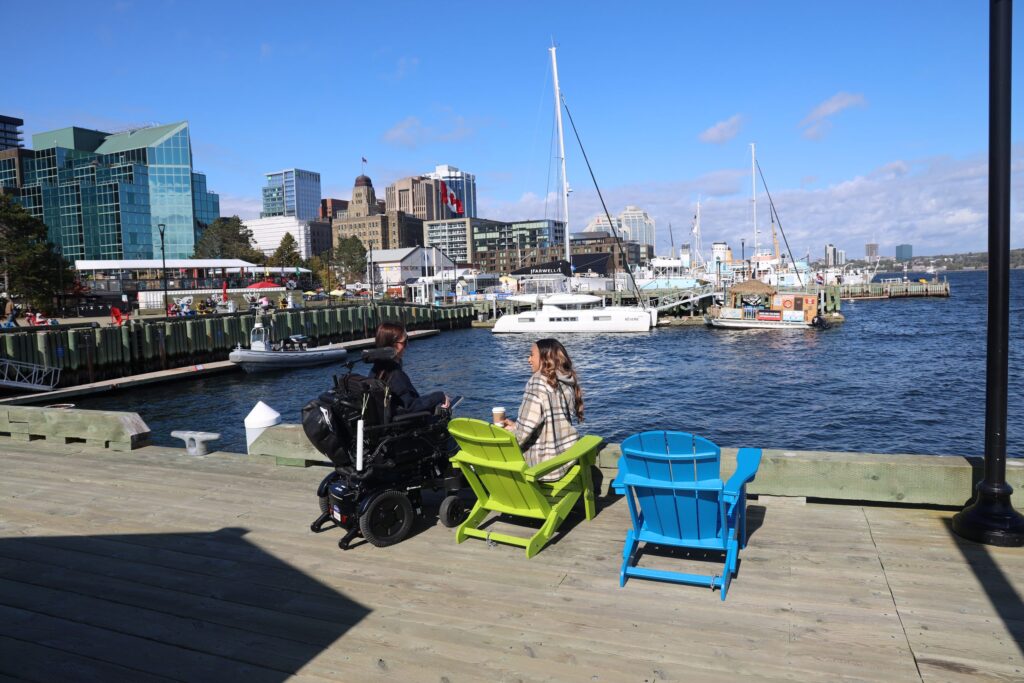 Tori Hunter, a wheelchair user and disabled travel blogger, sitting in her power wheelchair beside colourful Muskoka chairs on the Halifax waterfront boardwalk, looking out over Halifax Harbour and the downtown skyline in Nova Scotia, Canada