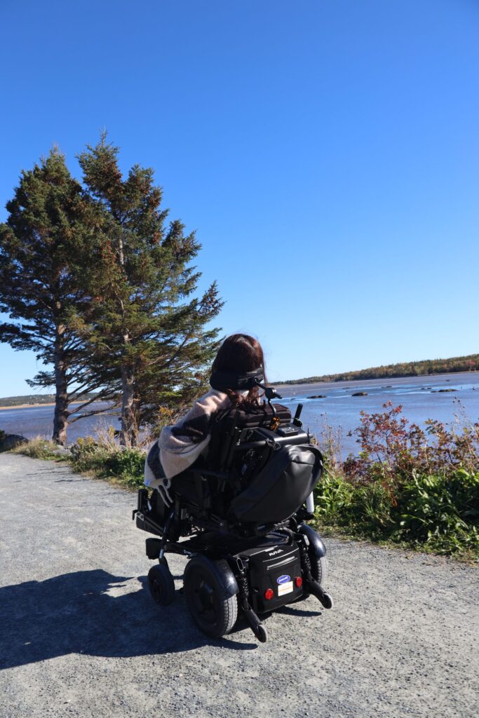 Tori Hunter, a wheelchair user and disabled travel blogger, rolling her power wheelchair along the level packed gravel causeway of the wheelchair accessible Salt Marsh Trail near Halifax, Nova Scotia, with the Cole Harbour salt marsh on both sides