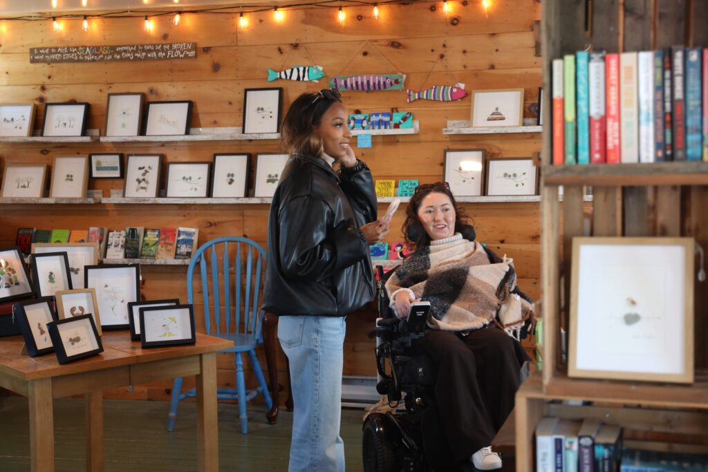 Tori Hunter, a wheelchair user and disabled travel blogger, browsing local sea glass art in her power wheelchair with a friend inside an artisan shop at Fisherman's Cove in Eastern Passage, Nova Scotia