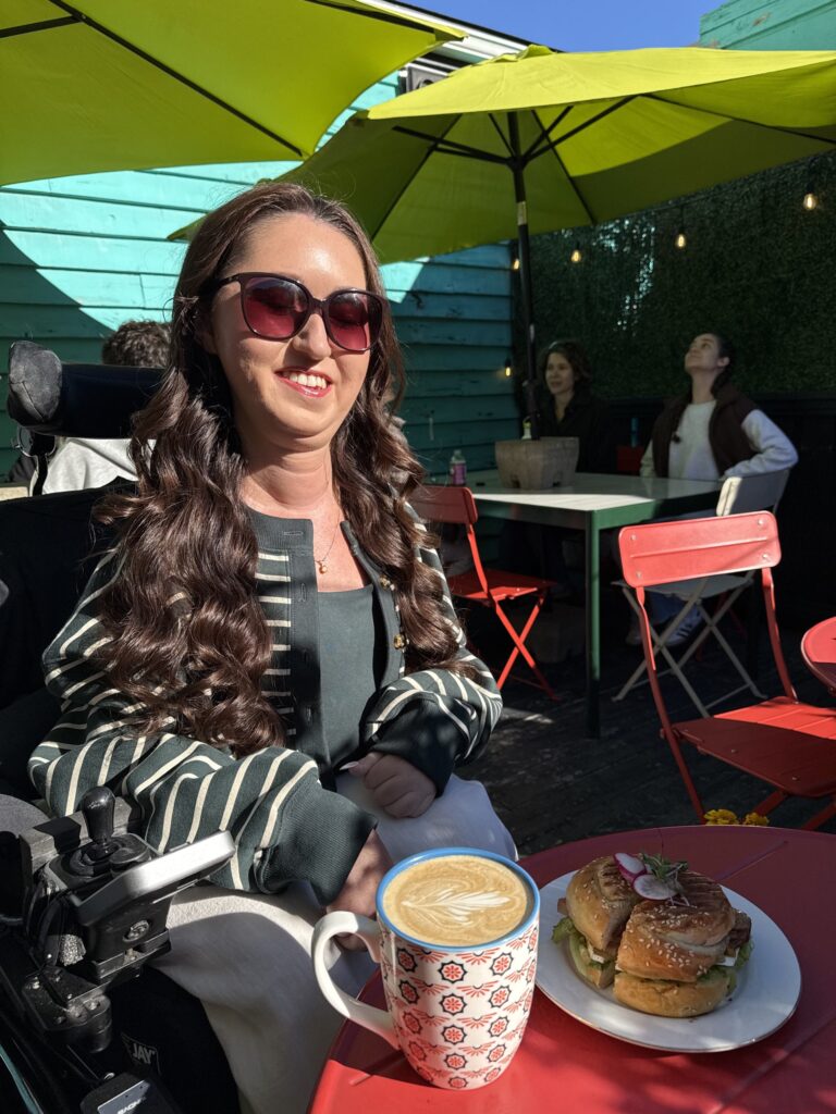 Tori Hunter, a wheelchair user and disabled travel blogger, smiling in her power wheelchair with a breakfast sandwich and a latte on the sunny patio at Dilly Dally Café, reached by a ramp, in Halifax, Nova Scotia