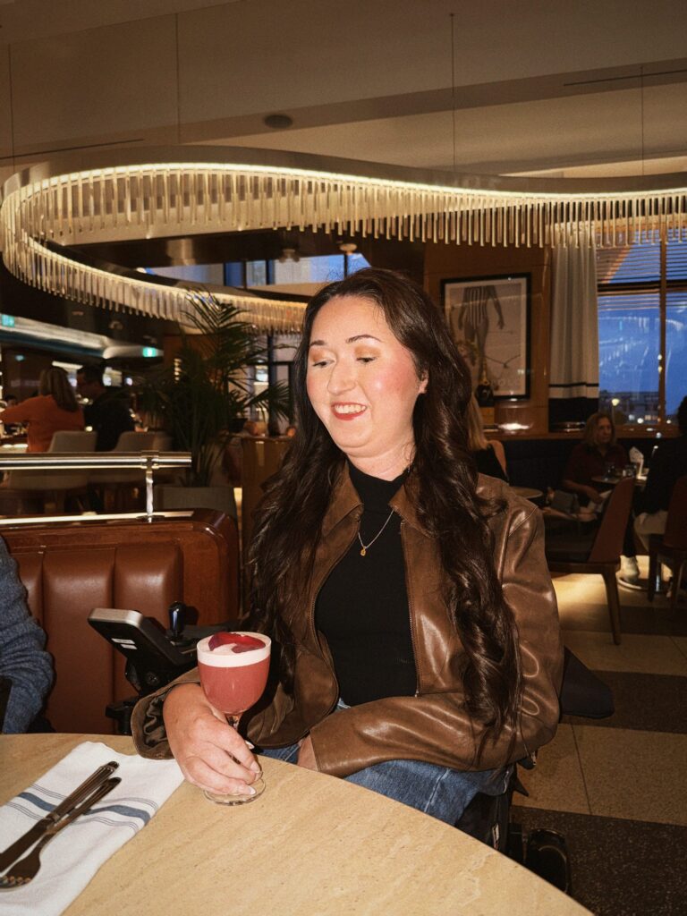 Tori Hunter, a wheelchair user and disabled travel blogger, smiling with a pink cocktail at her table in Waterpolo, a wheelchair accessible restaurant on the Halifax waterfront in Nova Scotia