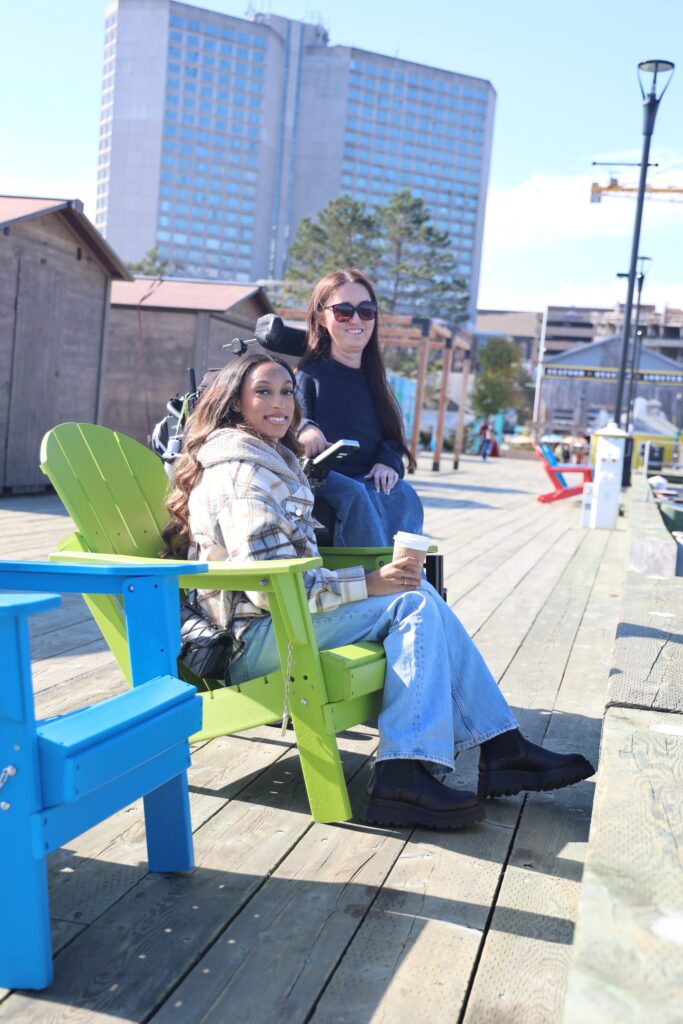 Tori Hunter, a wheelchair user and disabled travel blogger, smiling in her power wheelchair with a friend beside bright blue and green Muskoka chairs on the step-free Halifax waterfront boardwalk in Nova Scotia, Canada