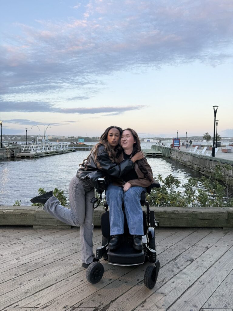 Tori Hunter, a wheelchair user and disabled travel blogger, hugging a friend in her power wheelchair on the Halifax waterfront boardwalk at sunset, with Halifax Harbour stretching out behind them in Nova Scotia, Canada