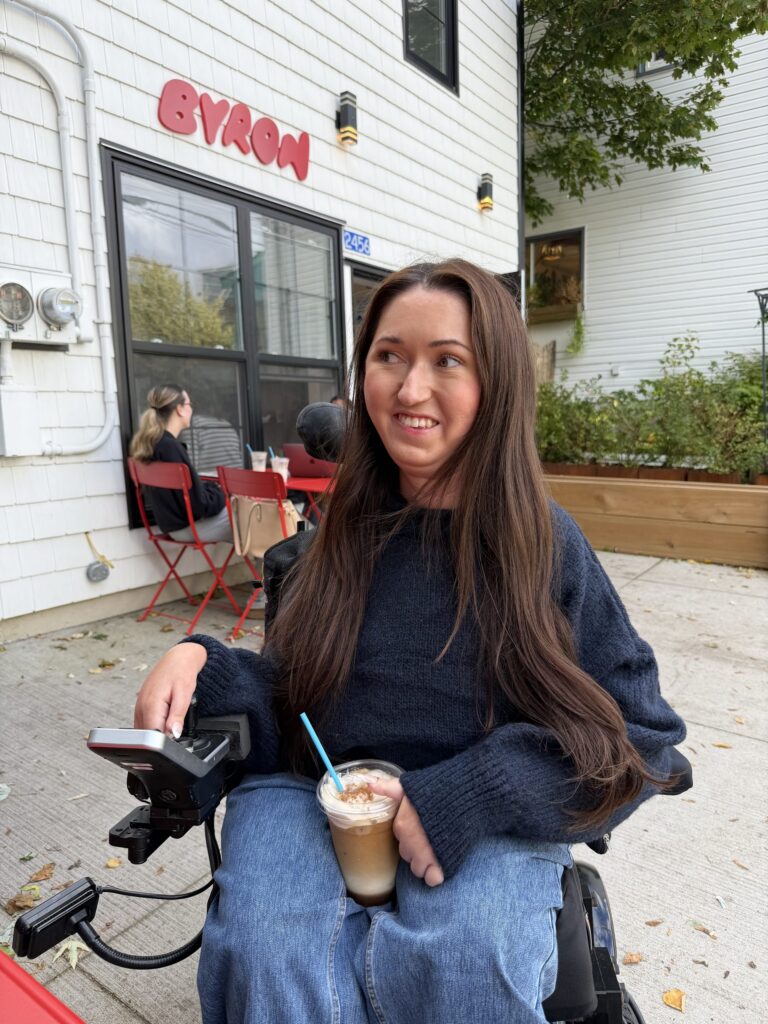 Tori Hunter, a wheelchair user and disabled travel blogger, smiling with an iced coffee in her power wheelchair outside the step-free entrance of Café Byron on Agricola Street in Halifax, Nova Scotia