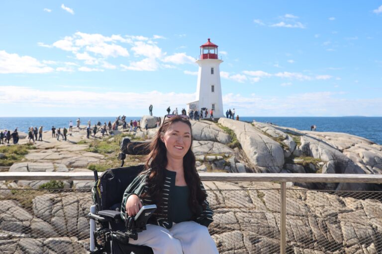 Tori Hunter, a wheelchair user and disabled travel blogger, smiling in her power wheelchair on the wheelchair accessible viewing platform at Peggy's Cove with the iconic Peggy's Point Lighthouse and granite rocks behind her in Nova Scotia, Canada