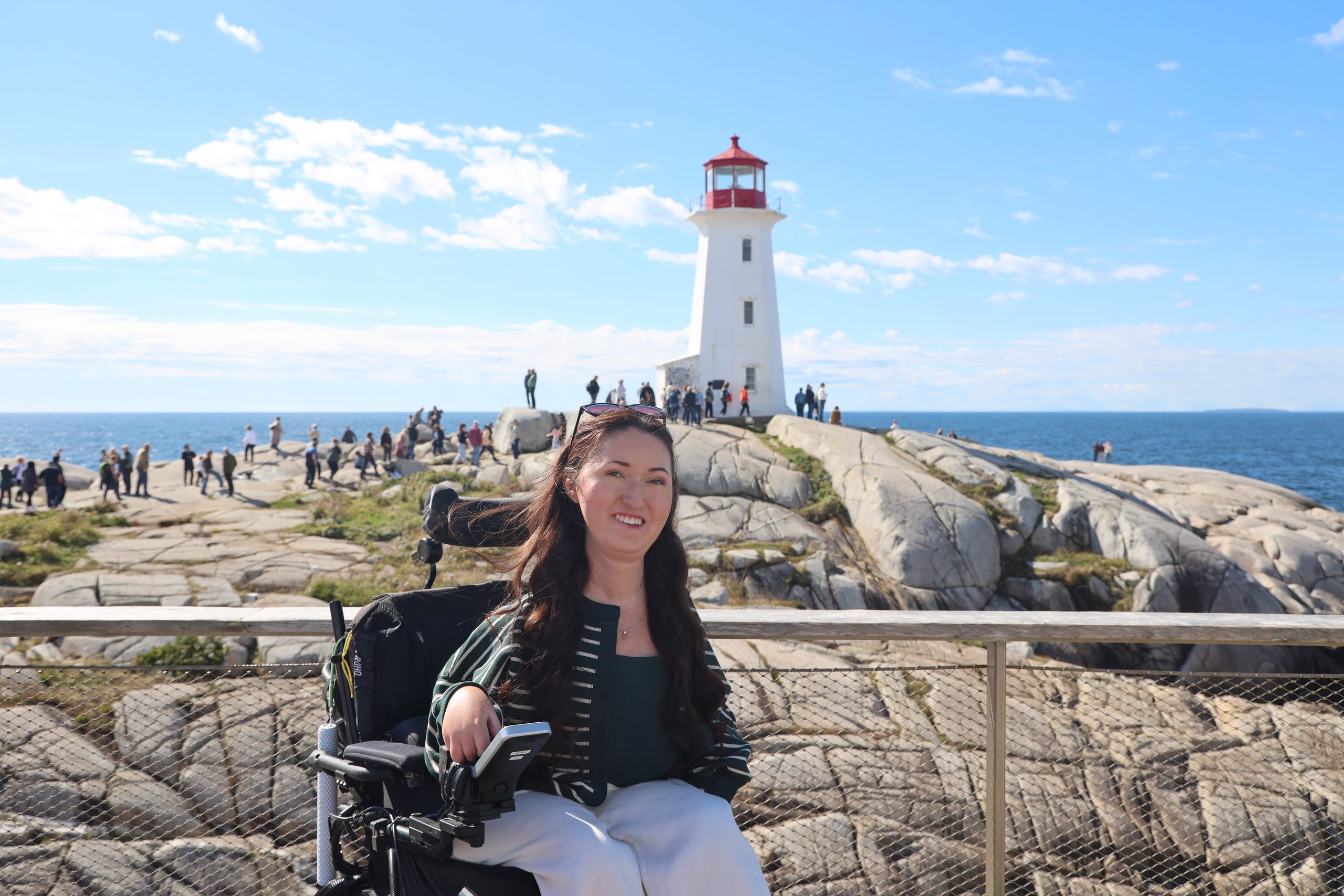 Tori Hunter, a wheelchair user and disabled travel blogger, smiling in her power wheelchair on the wheelchair accessible viewing platform at Peggy's Cove with the iconic Peggy's Point Lighthouse and granite rocks behind her in Nova Scotia, Canada