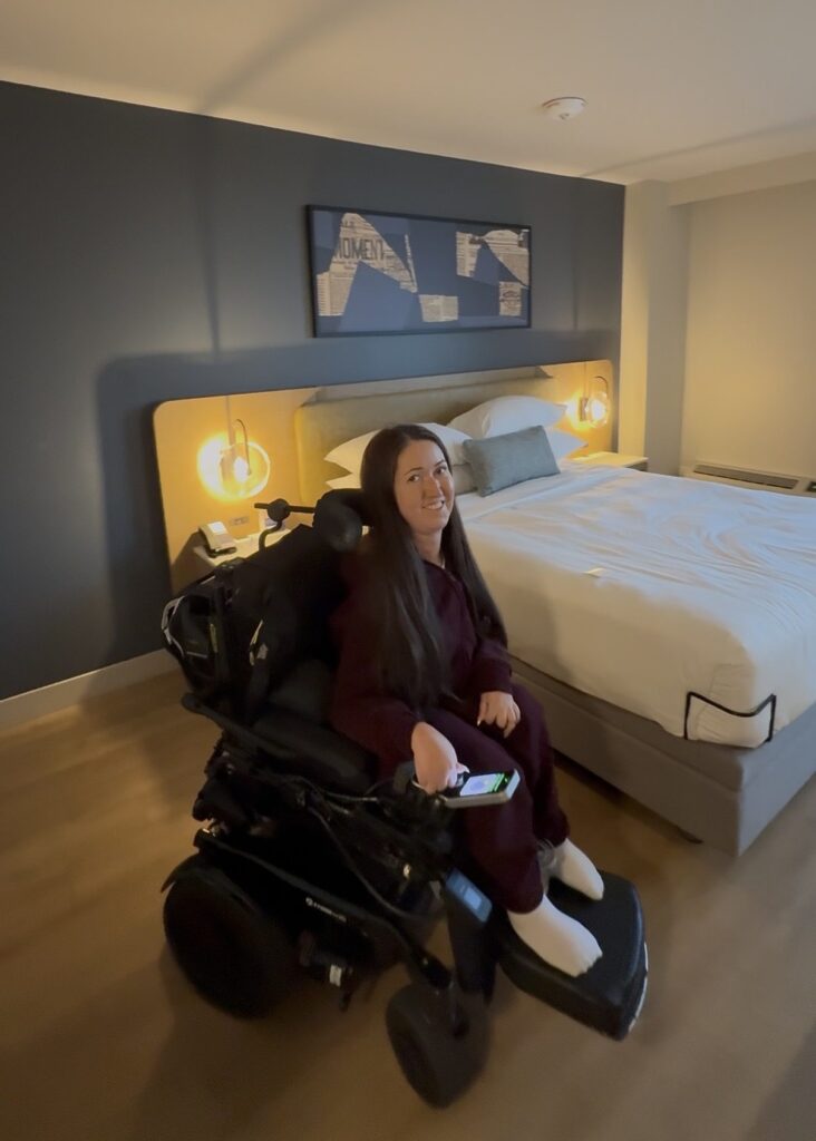 Tori Hunter, a wheelchair user and disabled travel blogger, smiling in her power wheelchair beside the bed in her accessible room at Delta Hotels Halifax, showing the clear floor space to manoeuvre a wheelchair