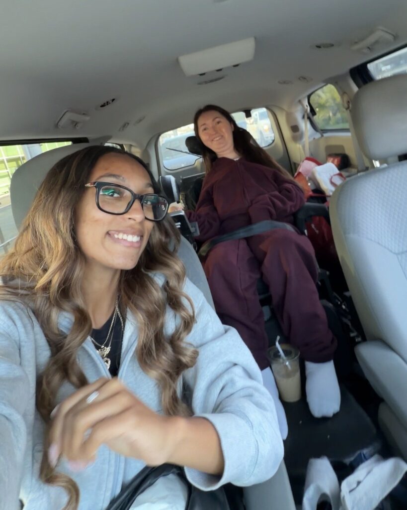 Tori Hunter, a wheelchair user and disabled travel blogger, smiling in her power wheelchair, secured with a seat belt in the back of a wheelchair accessible ExtraCare Taxi van, while her friend Alicia takes a selfie in Halifax, Nova Scotia