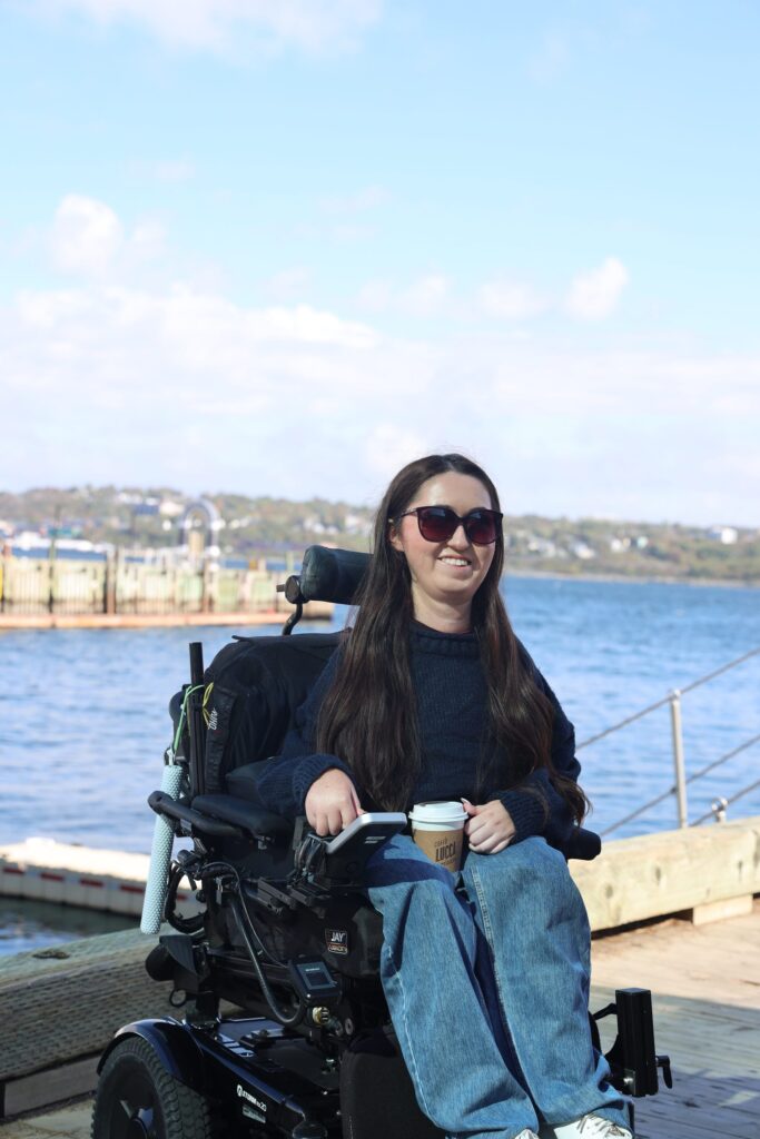 Tori Hunter, a wheelchair user and disabled travel blogger, smiling in her power wheelchair on the flat wooden Halifax waterfront boardwalk with Halifax Harbour behind her in Nova Scotia, Canada
