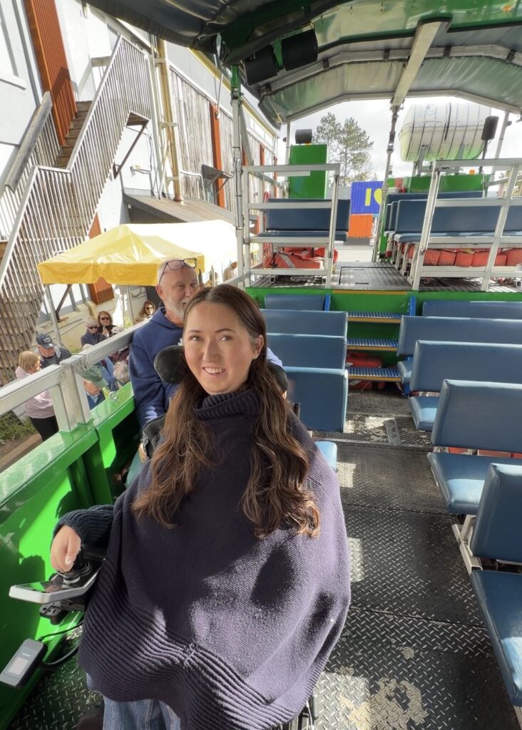 Tori Hunter, a wheelchair user and disabled travel blogger, smiling in her power wheelchair secured with tie-downs on board the wheelchair accessible Harbour Hopper tour in Halifax, Nova Scotia