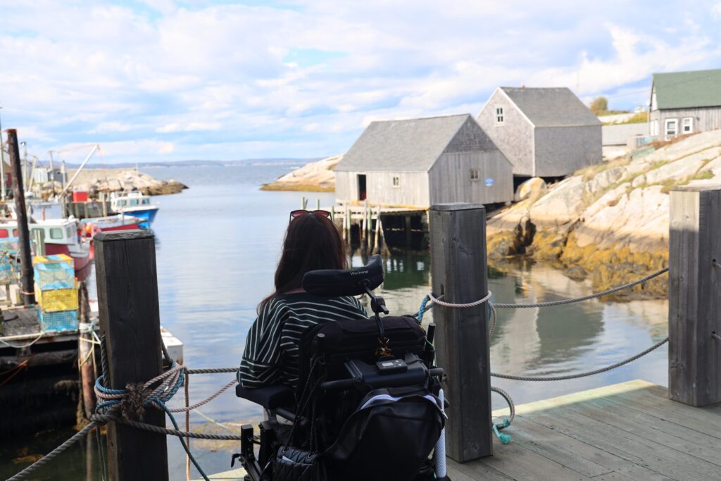 Tori Hunter, a wheelchair user and disabled travel blogger, in her power wheelchair on the wooden wharf looking out at the fishing shacks and boats in the harbour of Peggy's Cove village, Nova Scotia, Canada