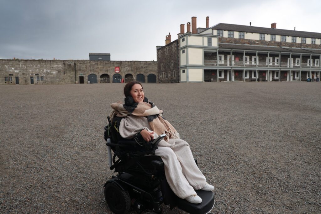 Tori Hunter, a wheelchair user and disabled travel blogger, smiling in her power wheelchair on the gravel parade ground of the Halifax Citadel National Historic Site, with the stone fort walls and barracks behind her in Nova Scotia, Canada