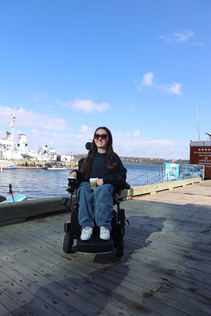 Tori Hunter, a wheelchair user and disabled travel blogger, rolling her power wheelchair along the wide, level wooden boardwalk on the wheelchair accessible Halifax waterfront in Nova Scotia, Canada