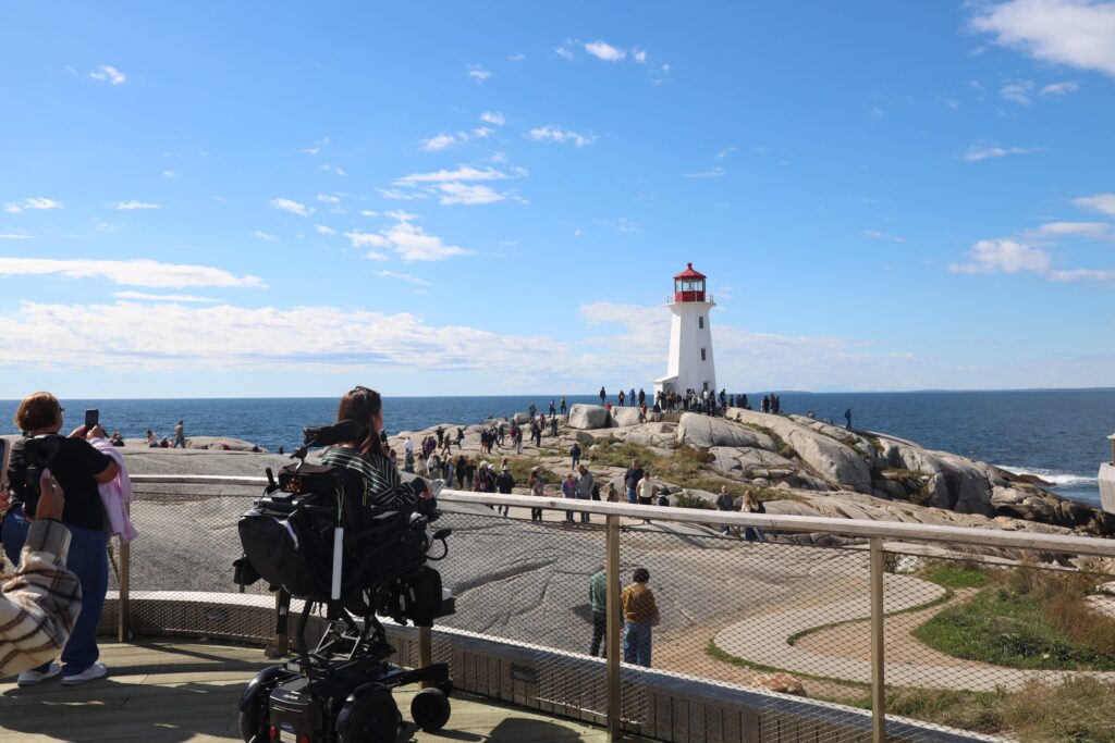 Tori Hunter, a wheelchair user and disabled travel blogger, in her power wheelchair on the Rick Hansen Gold certified accessible viewing platform at Peggy's Cove, looking out over the rocks to the lighthouse in Nova Scotia, Canada